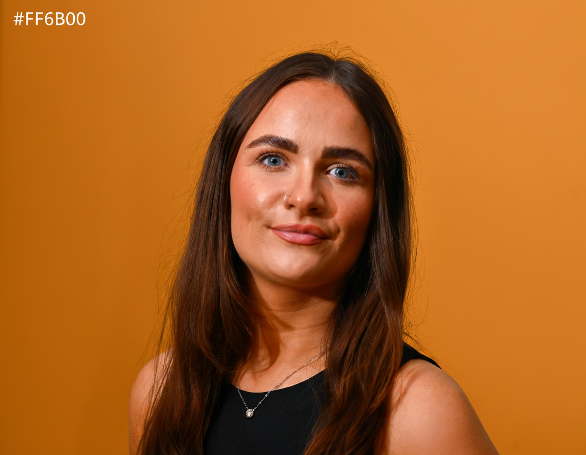 Take this portrait photo of Kirsty Adamson and replace the plain grey background with a bright orange (#FF6B00) background. Keep Kirsty exactly as she is — same pose, lighting, hair, face, clothing. Only change the background colour to solid bright orange.